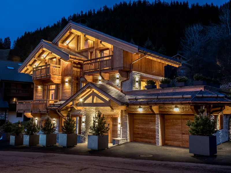 Chalet Arpège lit at dusk, snow on its balconies and lanterns along the approach, Méribel